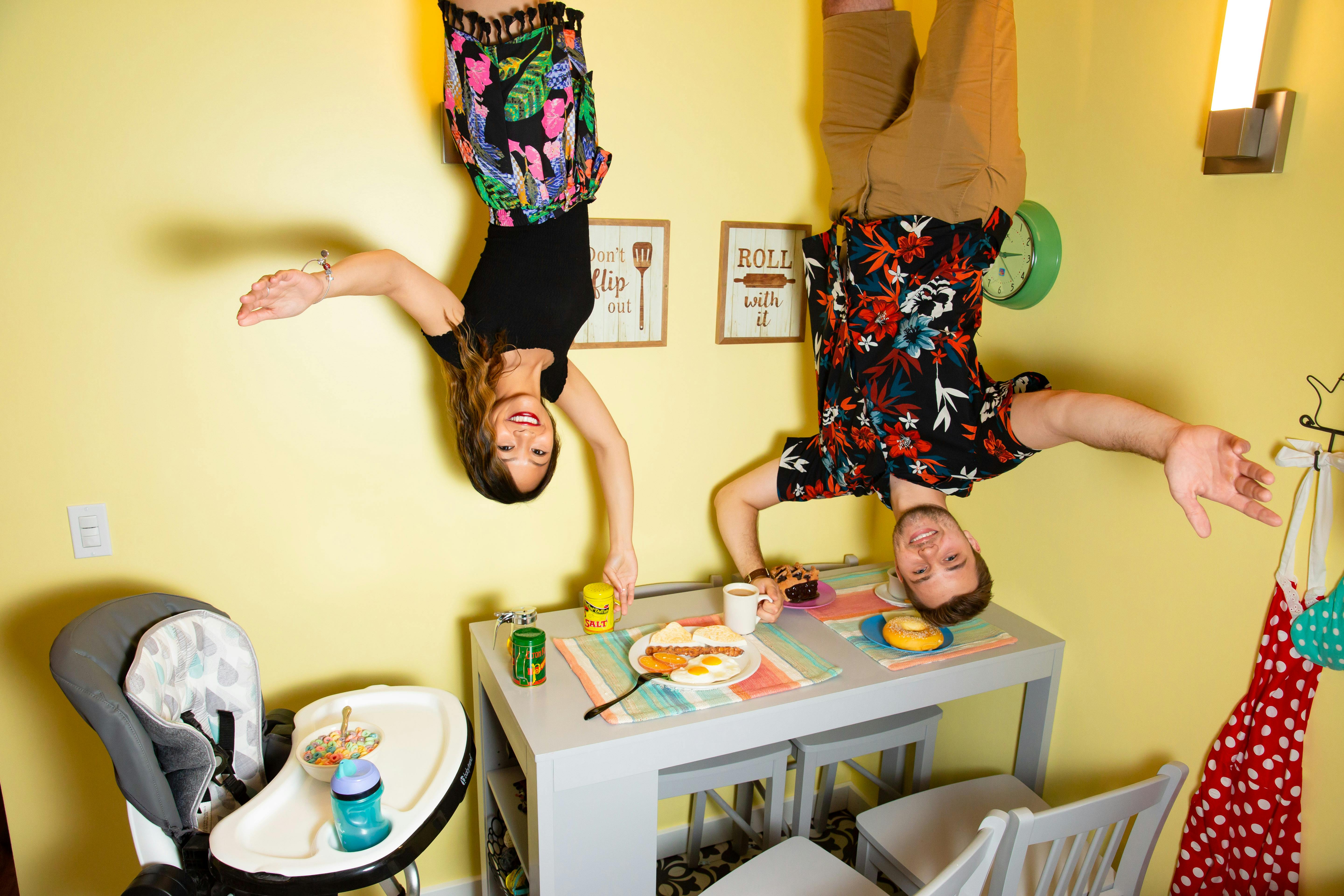Two people hanging upside down in a brightly colored kitchen with a dining table set for breakfast, creating an illusion.