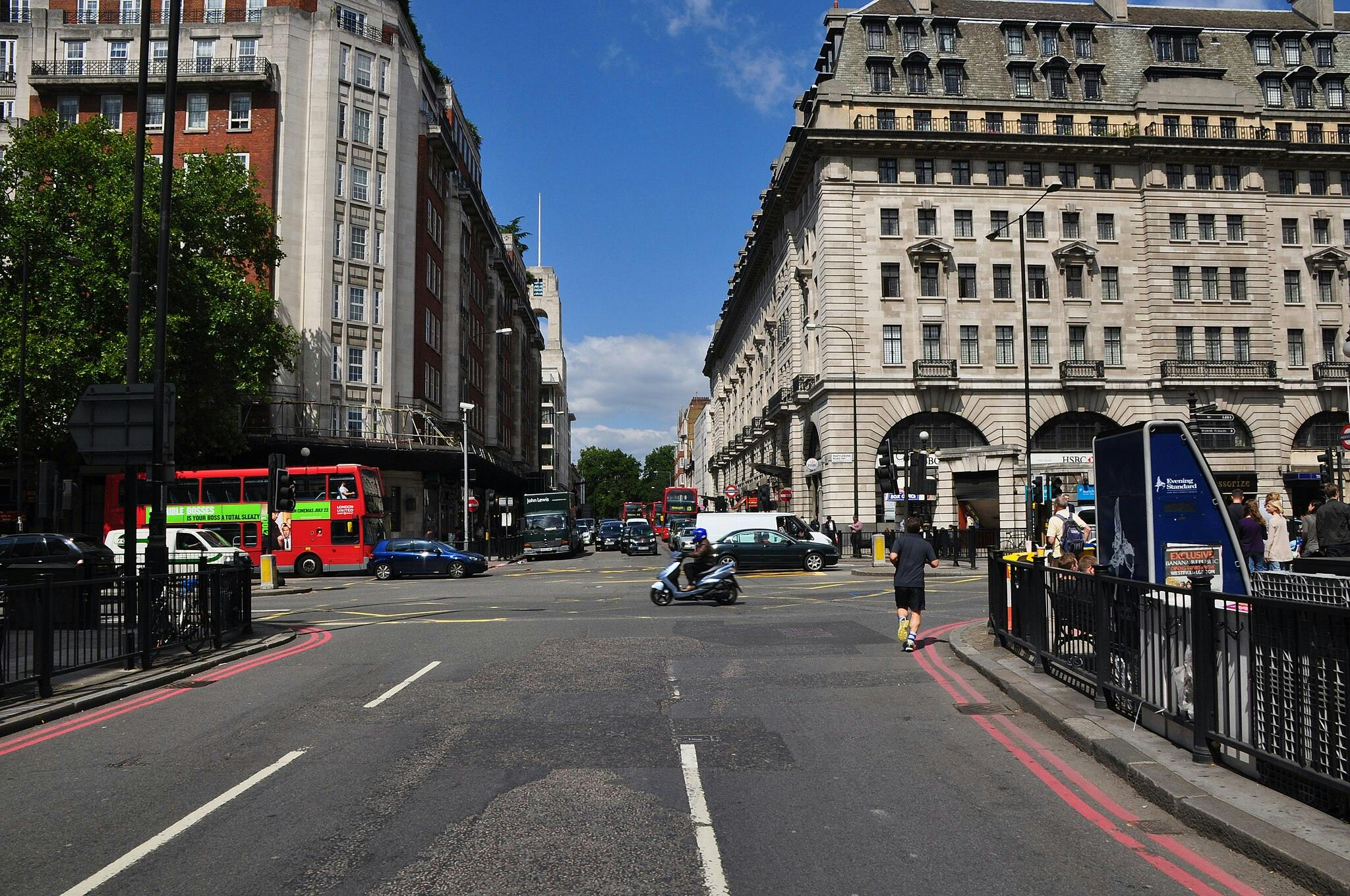 Bustling city intersection with pedestrians, vehicles, a red double-decker bus, and historic buildings under a blue sky.