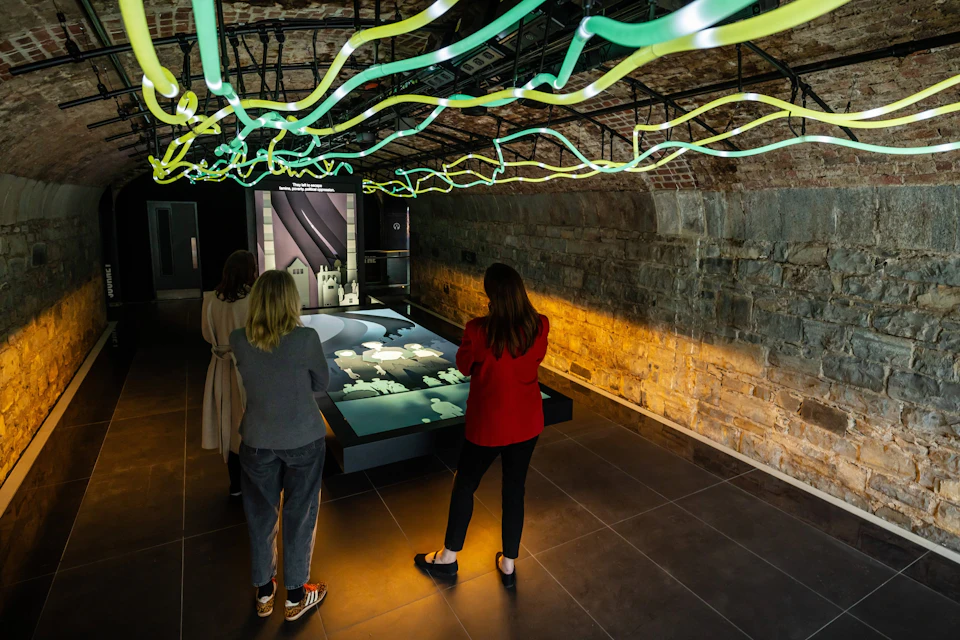 A group of people observing a modern art exhibit under a vaulted brick ceiling with neon light installations.