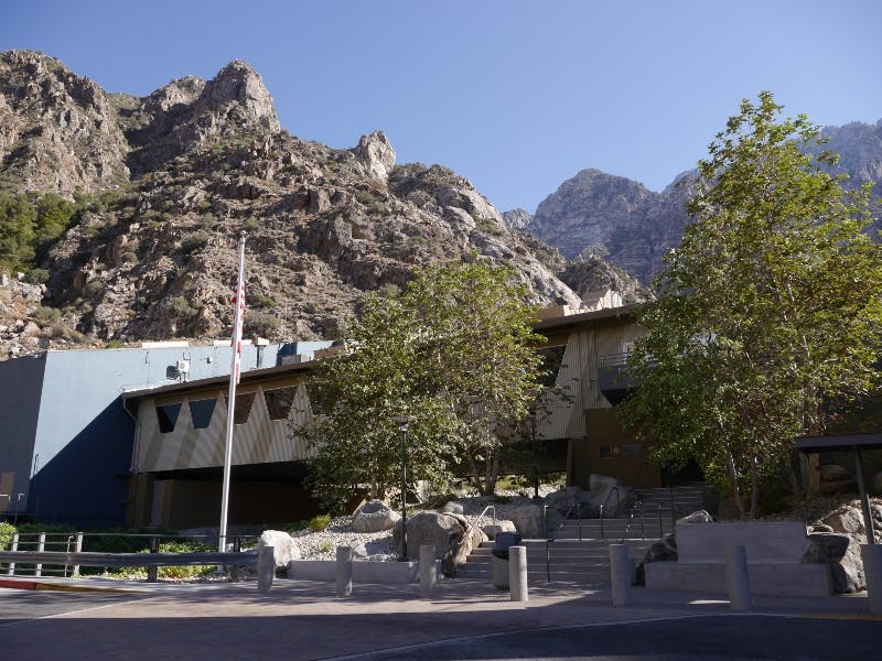 A modern building with a flagpole in front, surrounded by rocks and trees, set against mountainous terrain under a clear sky.