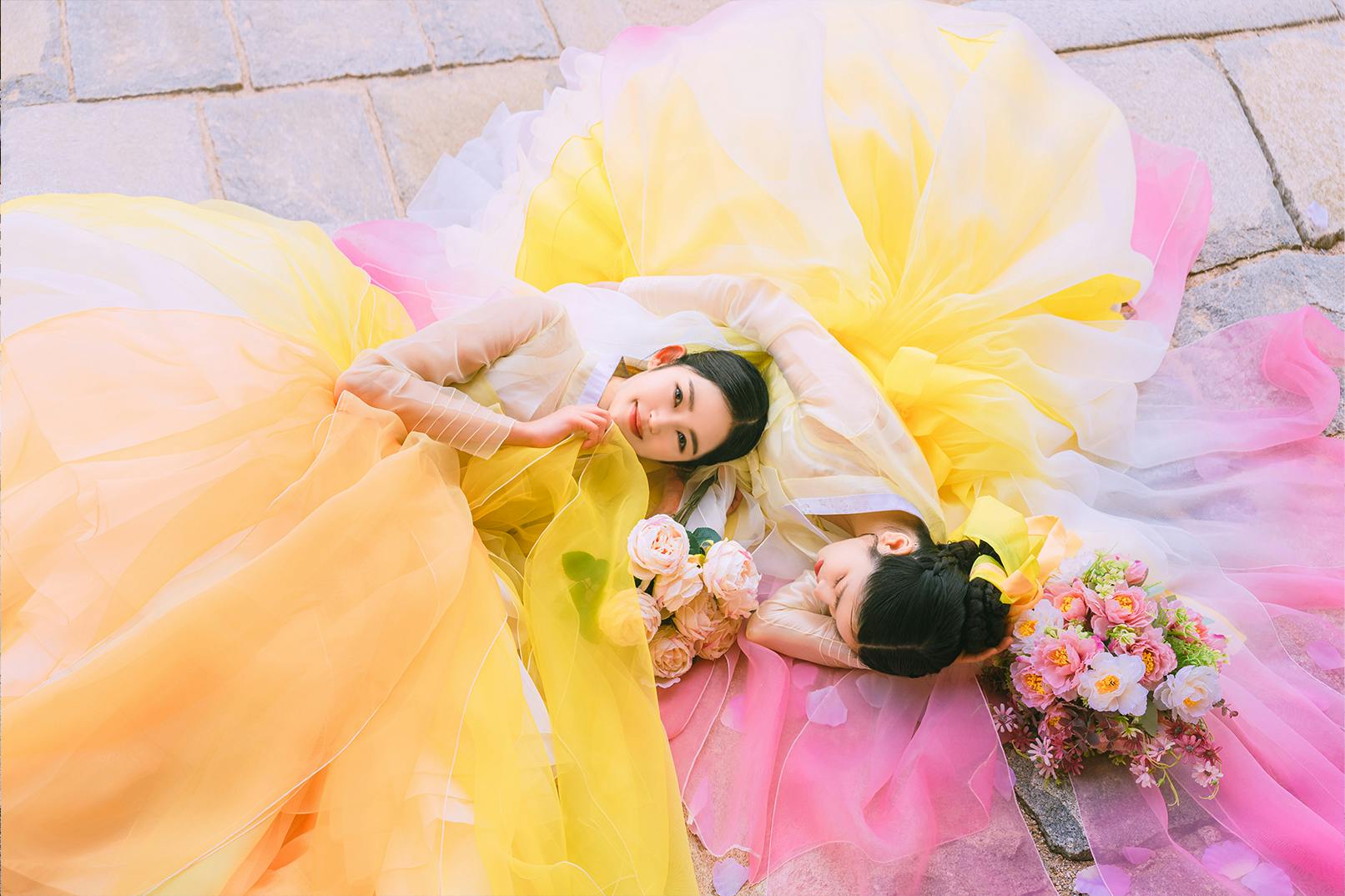 Two individuals in colorful flowing dresses, one yellow and one pink, lying on the ground holding flower bouquets.
