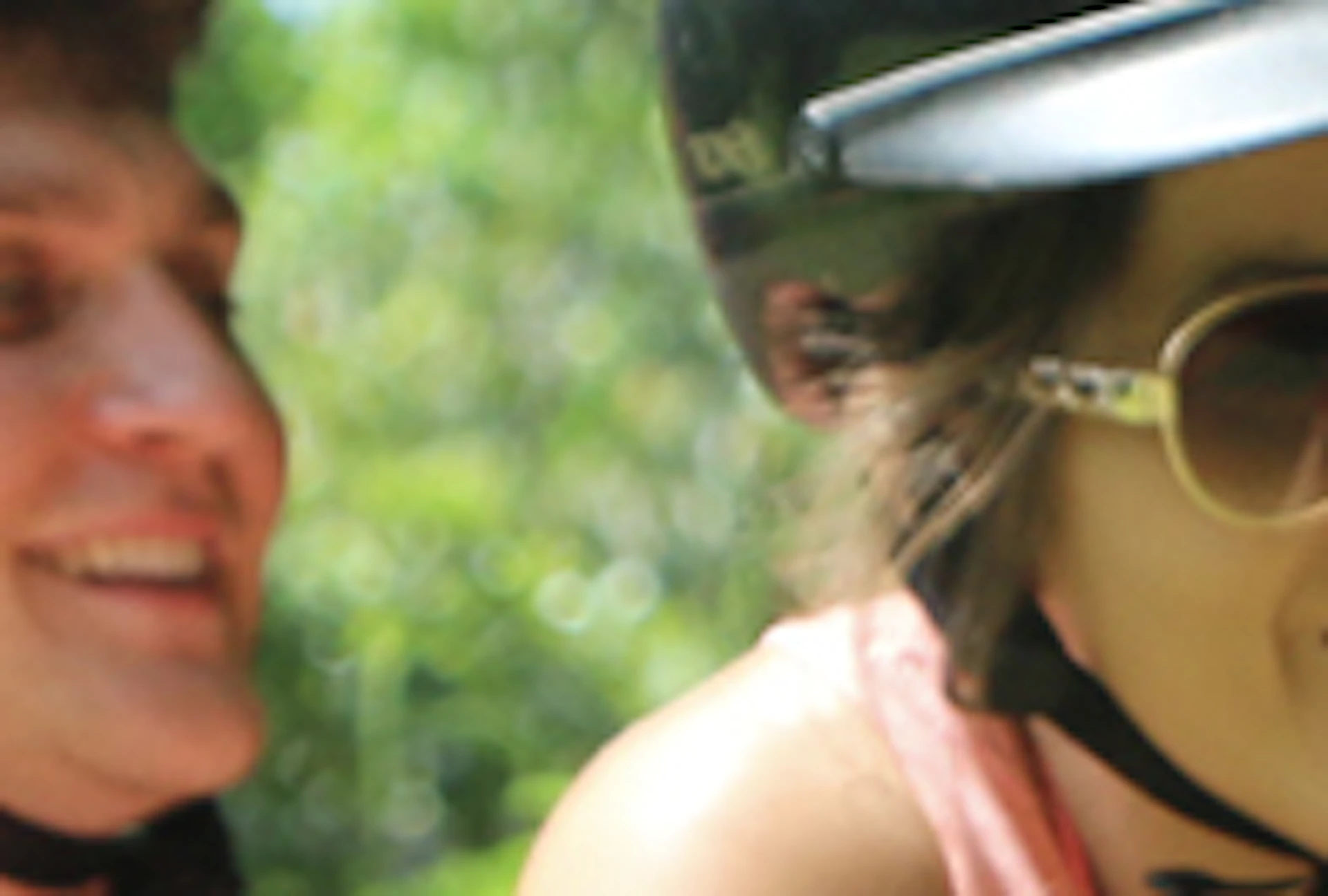 Two people wearing helmets and casual clothing ride a camel in a lush, green forested area.