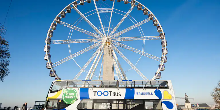 Un Tootbus de dos pisos amb la marca "Planet Friendly" està aparcat al davant d'una gran noria a Brussel·les sota un cel blau clar.
