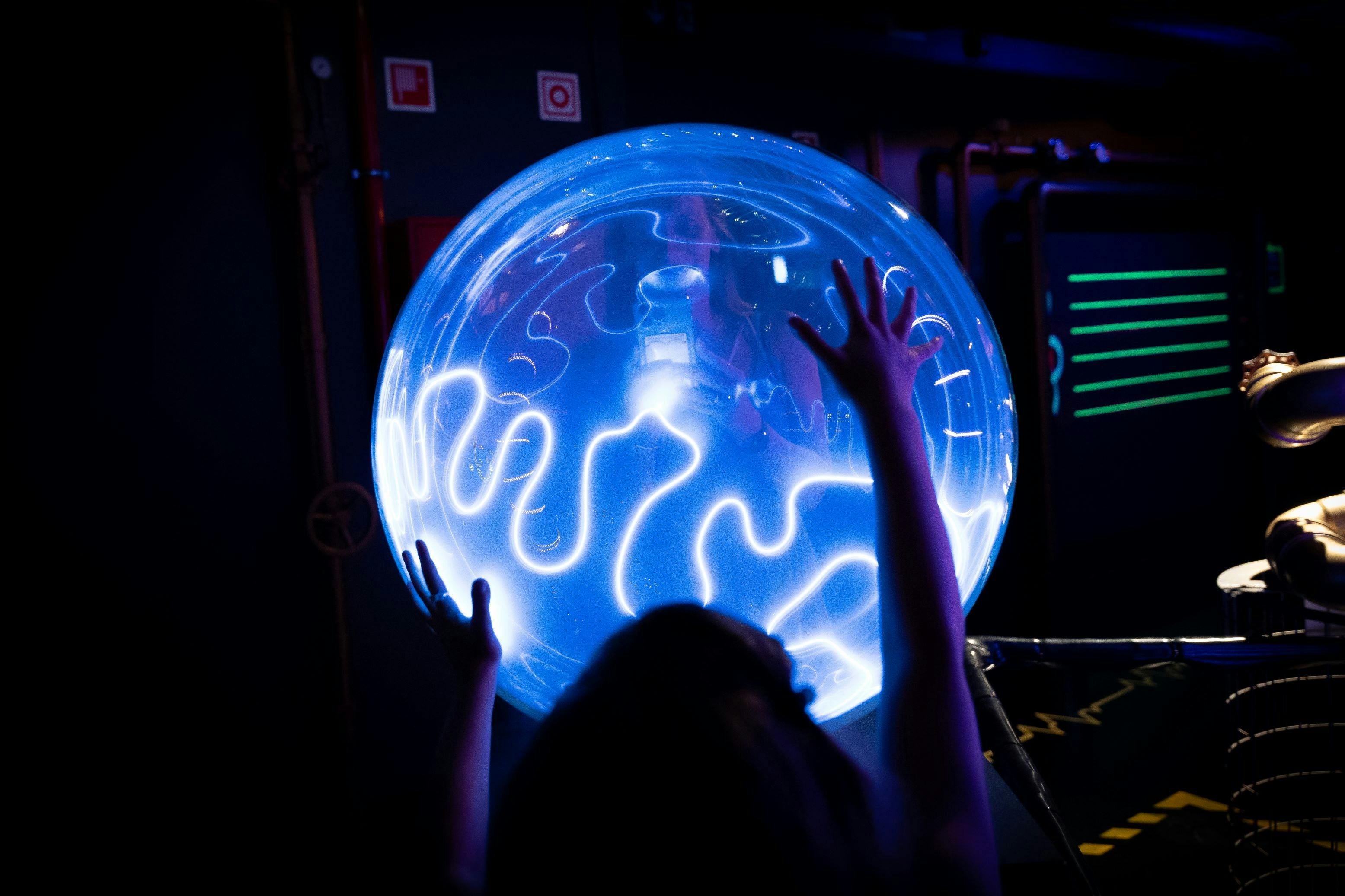 Person touching a glowing blue orb with hands against a dimly lit backdrop featuring pipes and control panels.