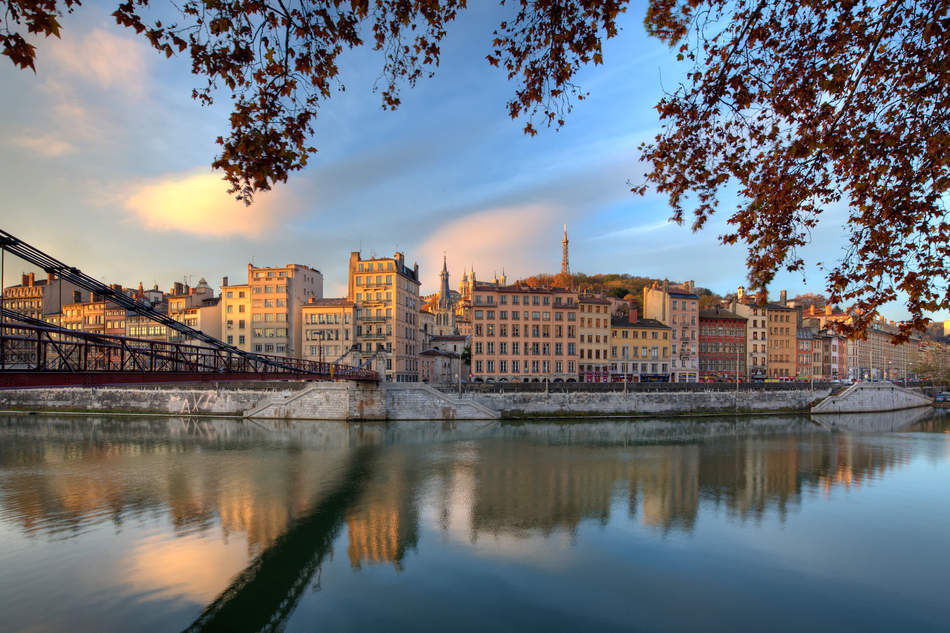 A riverside view of a city with historic buildings, a pedestrian bridge, and autumn trees under a blue and orange sky.
