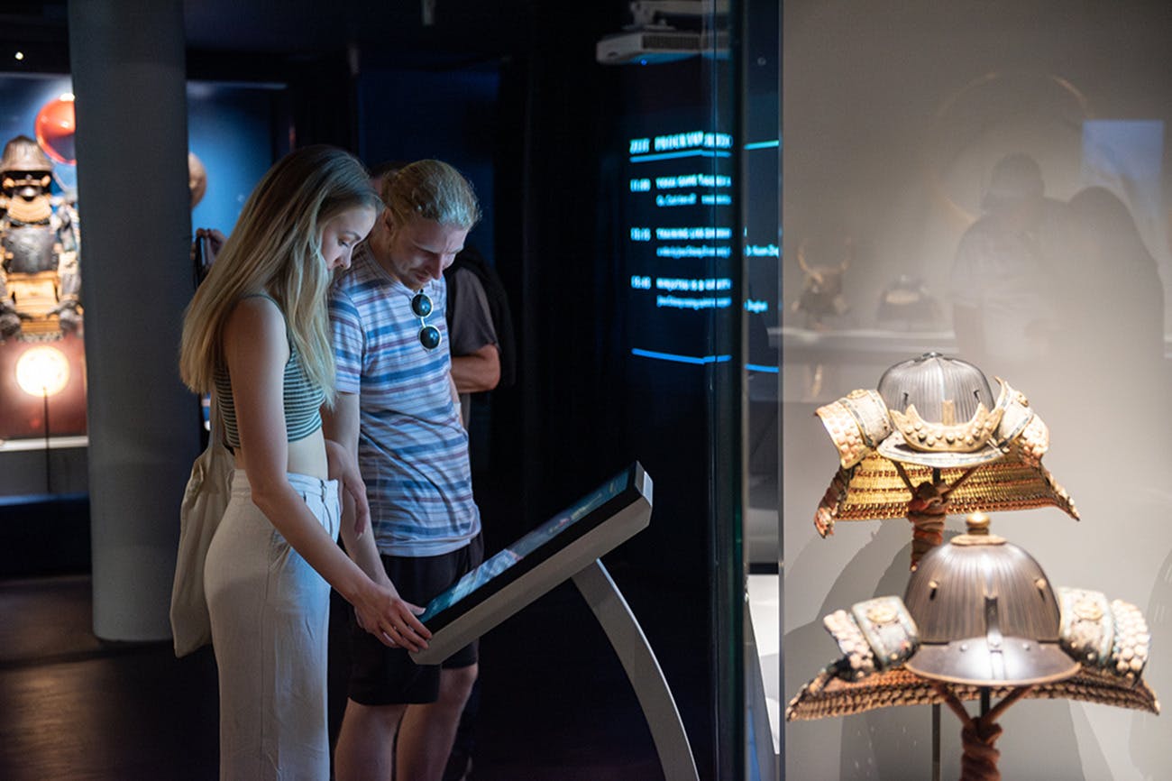 薄暗い博物館では、2人がタッチスクリーンディスプレイを操作し、近くのガラスケースには華やかなアーティファクトが見えます。