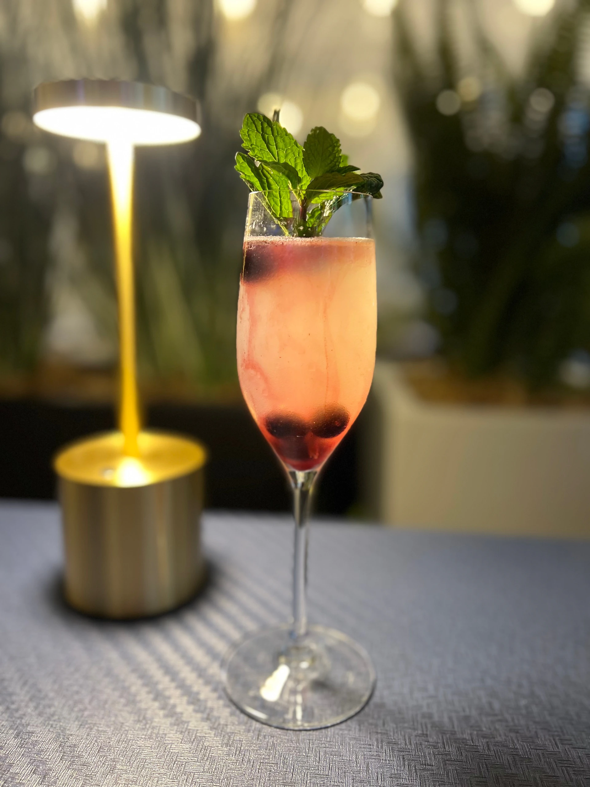 A pink cocktail in a flute glass garnished with mint leaves and berries, placed on a table near a warm-lit lamp.