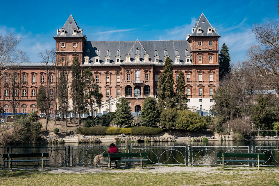 A person sits on a bench near a pond with a large historic building and greenery in the background under a clear blue sky.