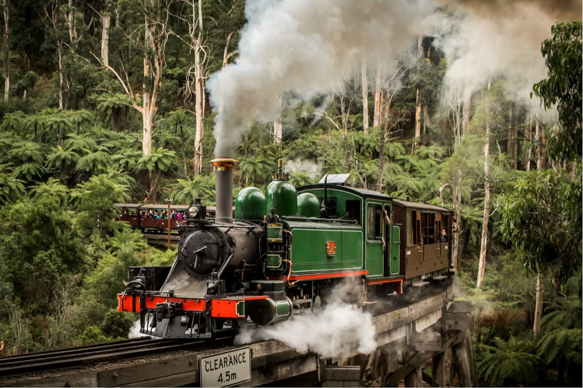 A green steam locomotive emitting smoke crosses a wooden bridge surrounded by dense forest. Sign reads "Clearance 4.5m."