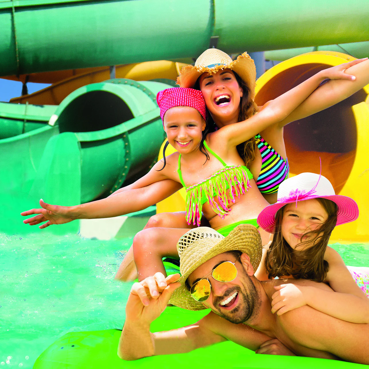 A joyful family of four in swimwear, wearing hats, at a water park with colorful slides in the background.