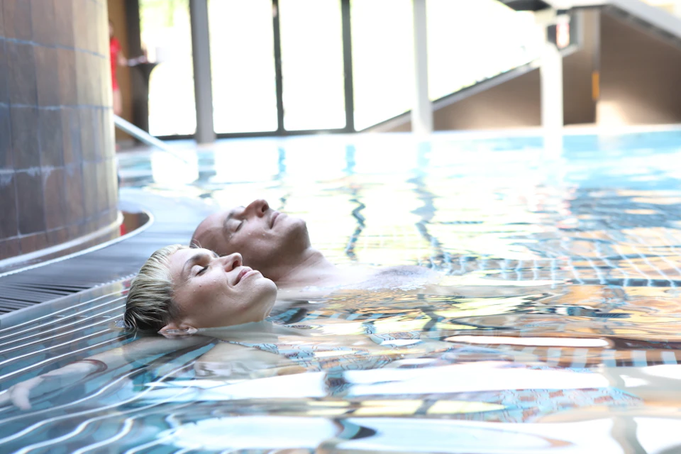 Two people floating on their backs in an indoor pool, eyes closed, with large windows in the background.