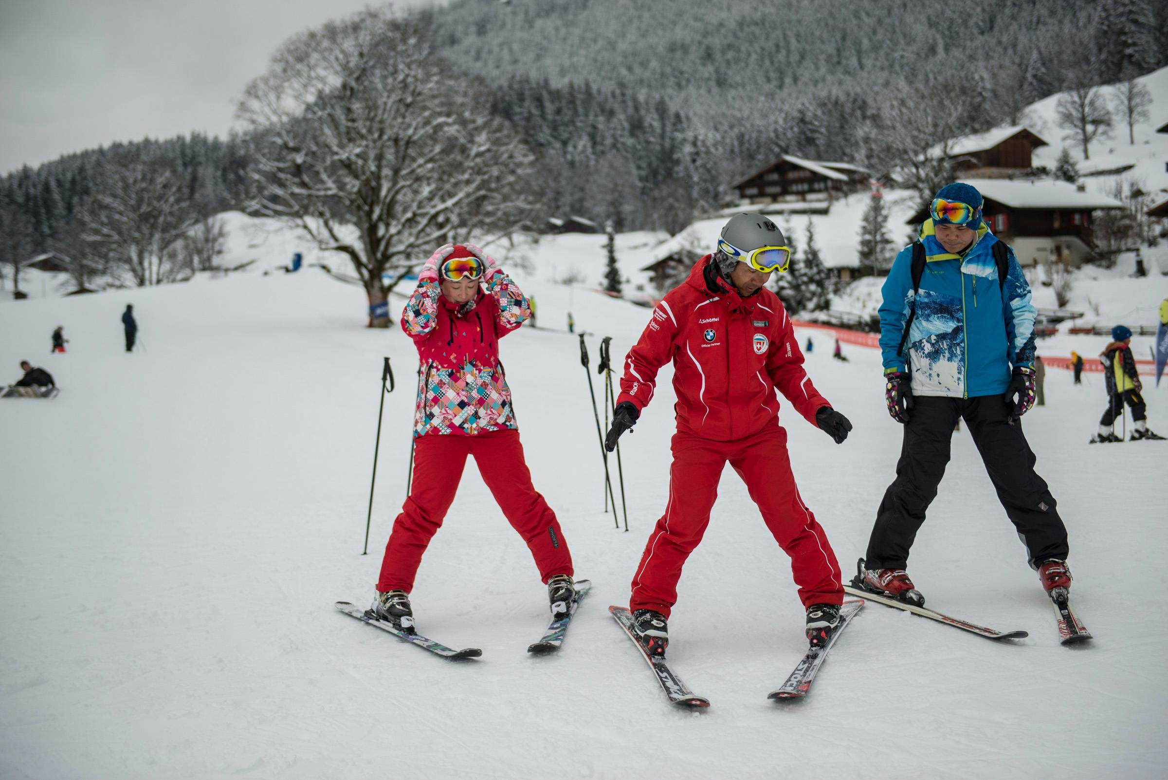 Δύο σκιέρ, ένας με πολύχρωμο μπουφάν και ένας με κόκκινο, προετοιμάζονται σε μια χιονισμένη πλαγιά με δέντρα και σπίτια στο παρασκήνιο.