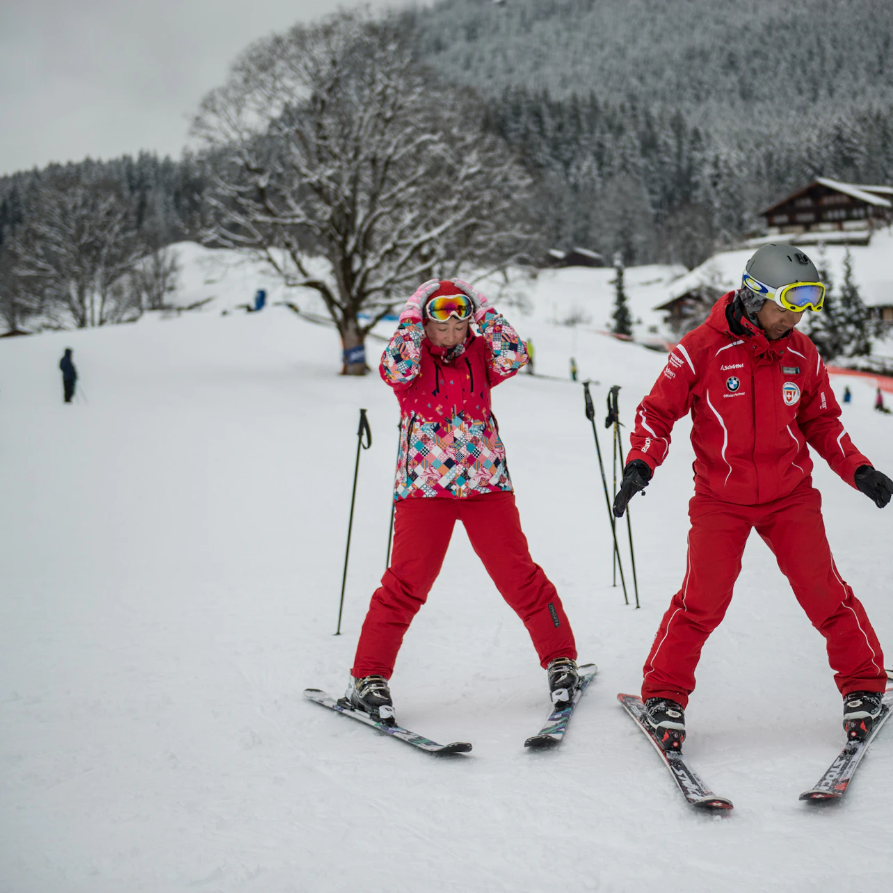 Two skiers, one in a colorful jacket and one in red, prepare on a snowy slope with trees and houses in the background.