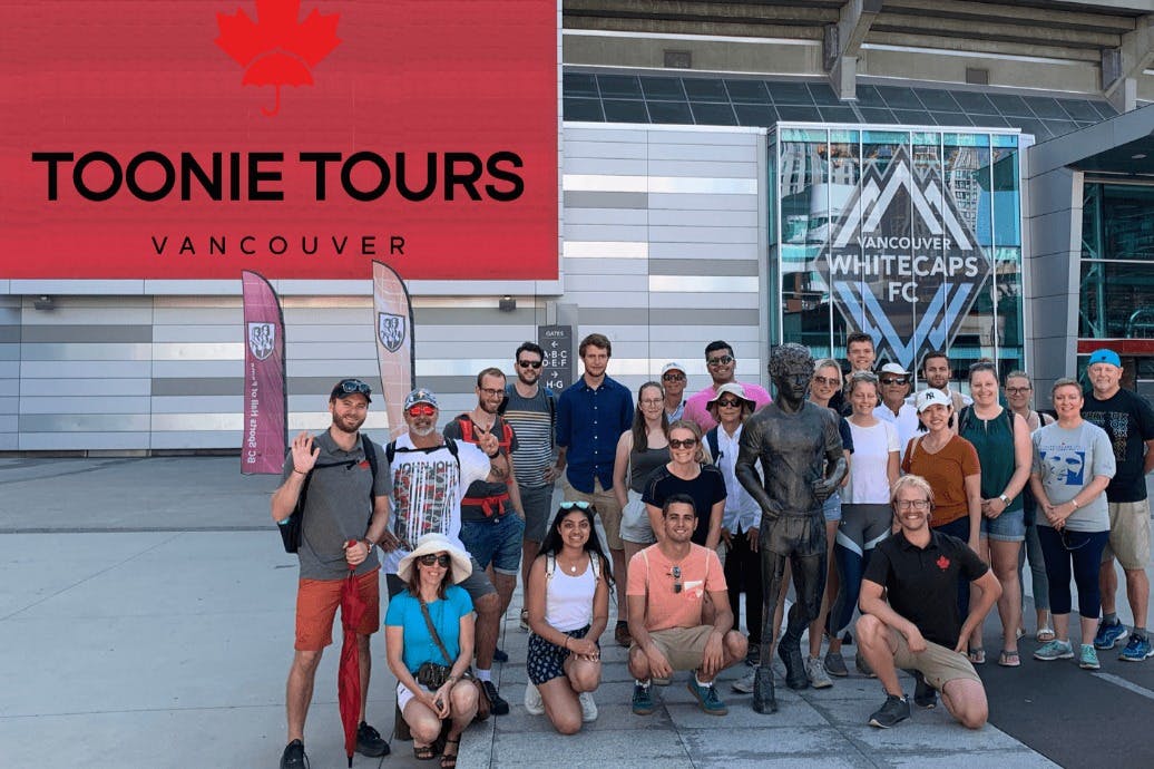 Skupina lidí pózuje kolem sochy v městském prostředí. Za nimi jsou vidět značky na Vancouver Whitecaps FC.