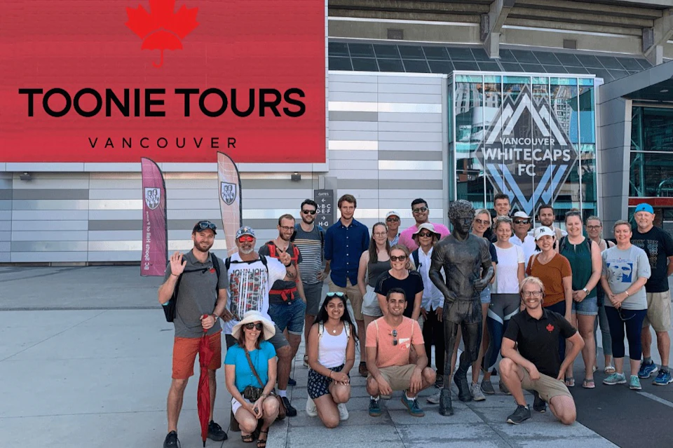 A group of people poses around a statue in an urban setting. Behind them, signs for Vancouver Whitecaps FC are visible.
