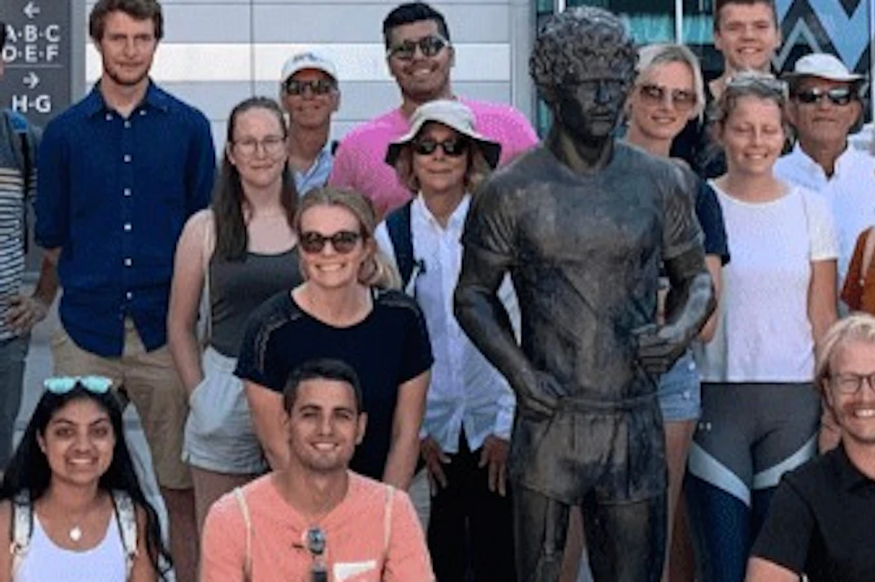 A group of people poses around a statue in an urban setting. Behind them, signs for Vancouver Whitecaps FC are visible.