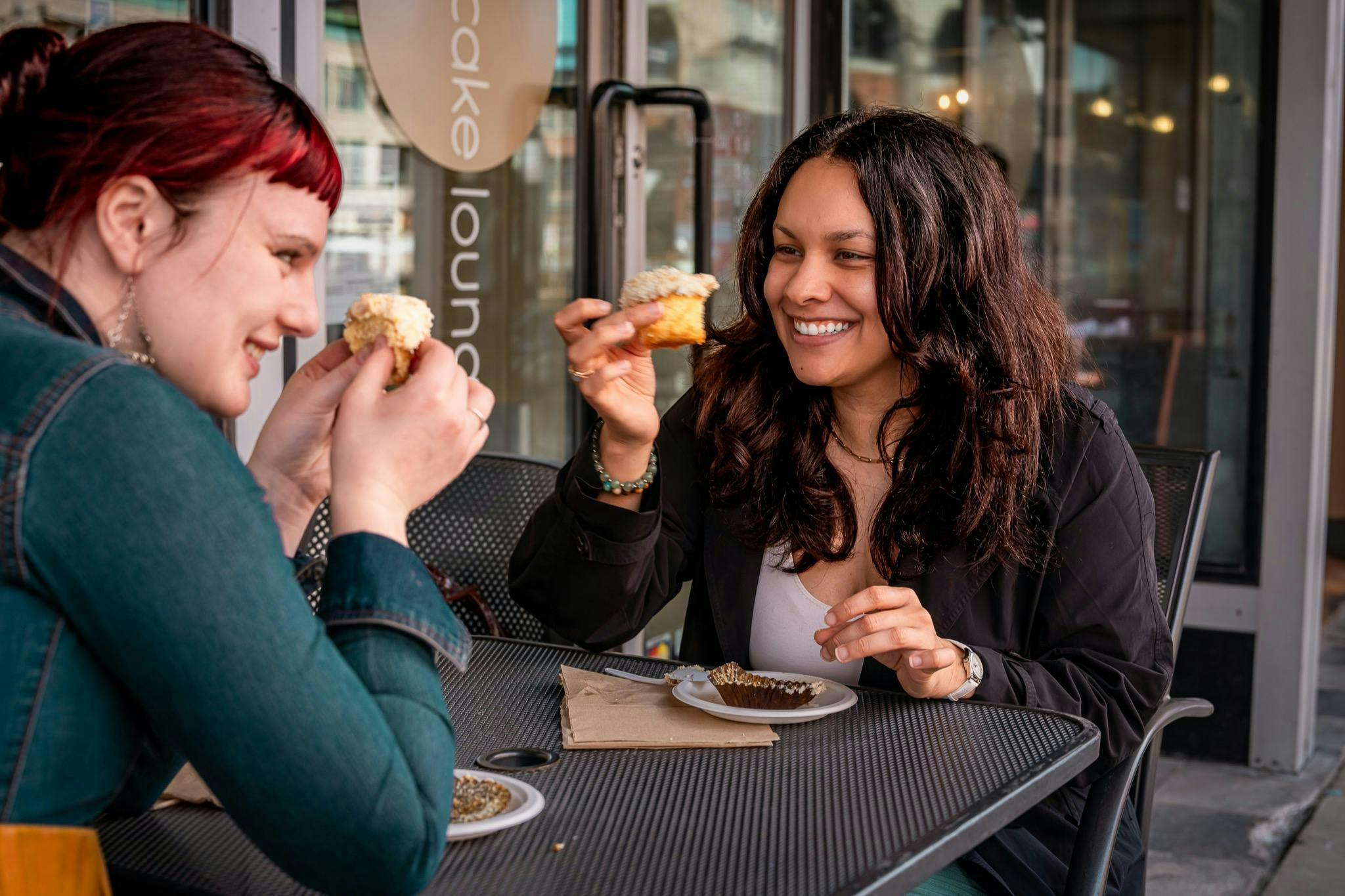 Una donna che sorride mentre tiene in mano un cupcake al tavolo di un bar all'aperto e si confronta con un'altra persona che tiene in mano un cupcake.