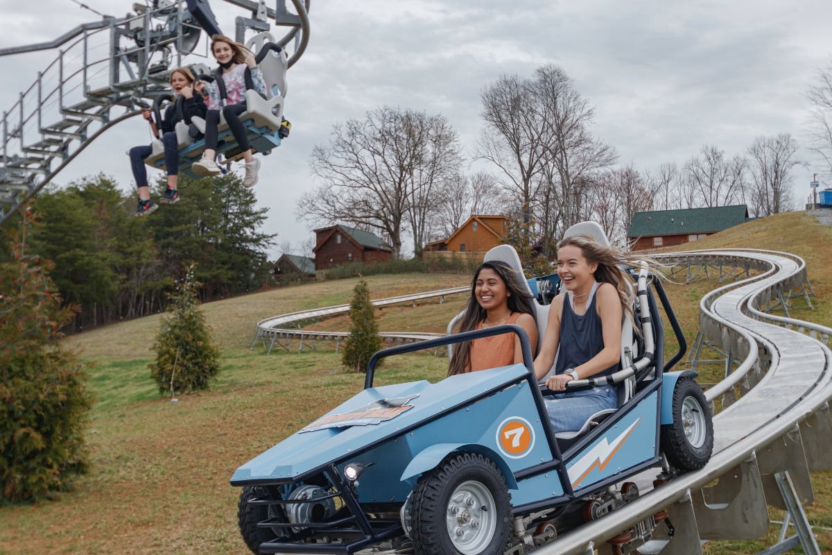 People ride a blue cart and seats on a hillside rail, with trees and wooden cabins in the background. Two girls smile in the cart.