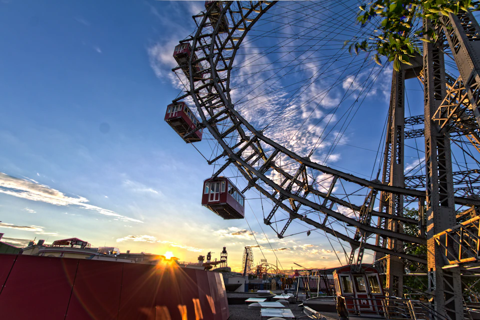A giant Ferris wheel with red gondolas against a sunset sky, with buildings and trees in the background.
