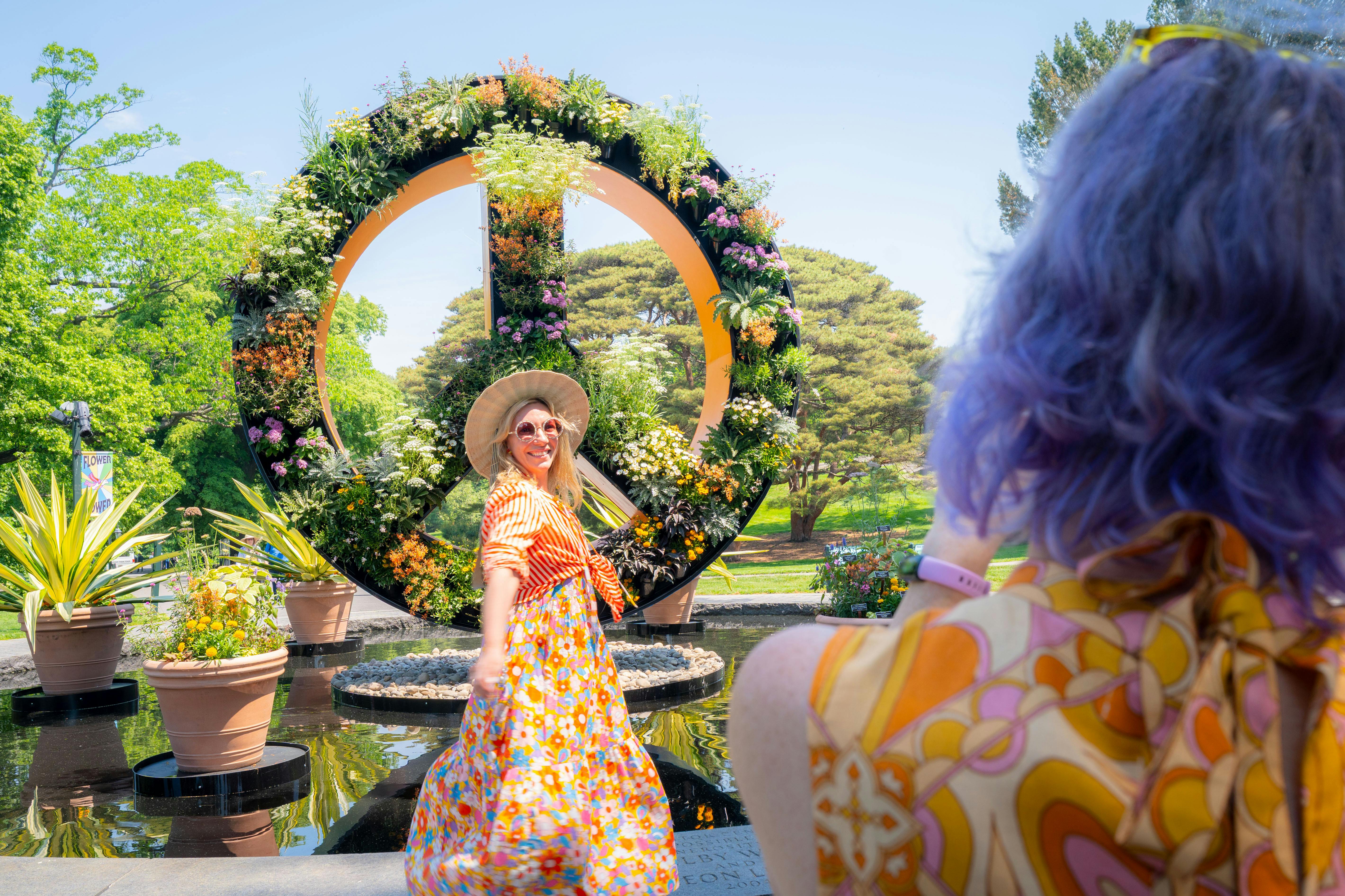 Una dona amb un vestit floral de colors i un barret posa davant d'un gran símbol de la pau adornat amb flors en un jardí.