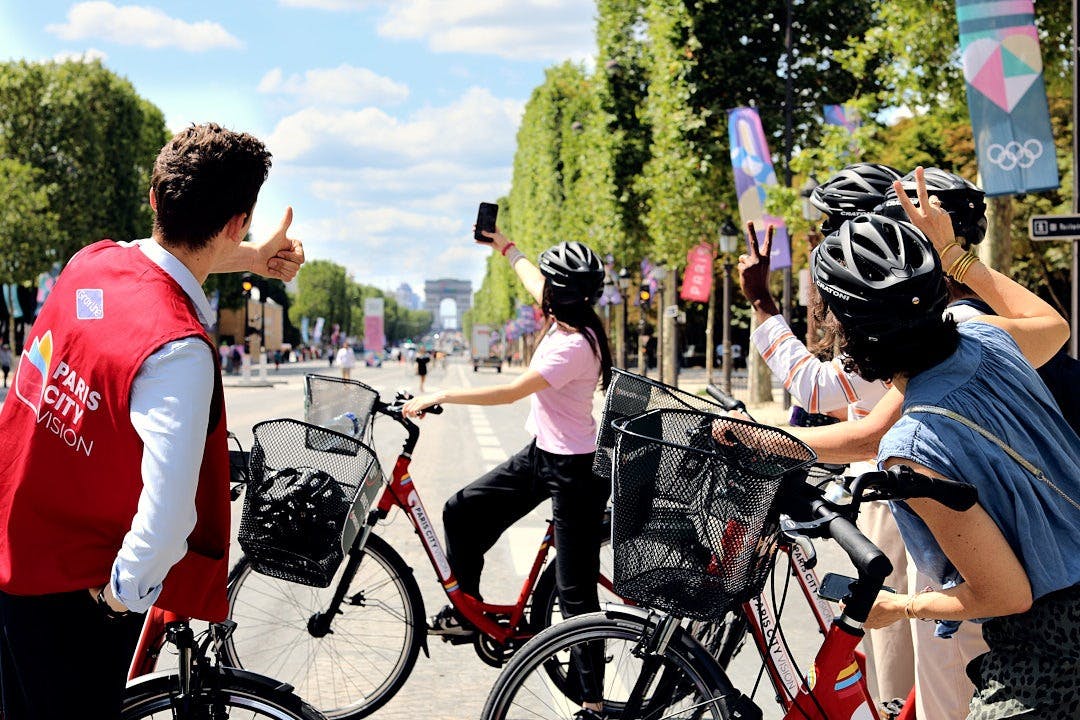 Group of people with bicycles posing and taking pictures on a tree-lined avenue with the Arc de Triomphe in the distance.