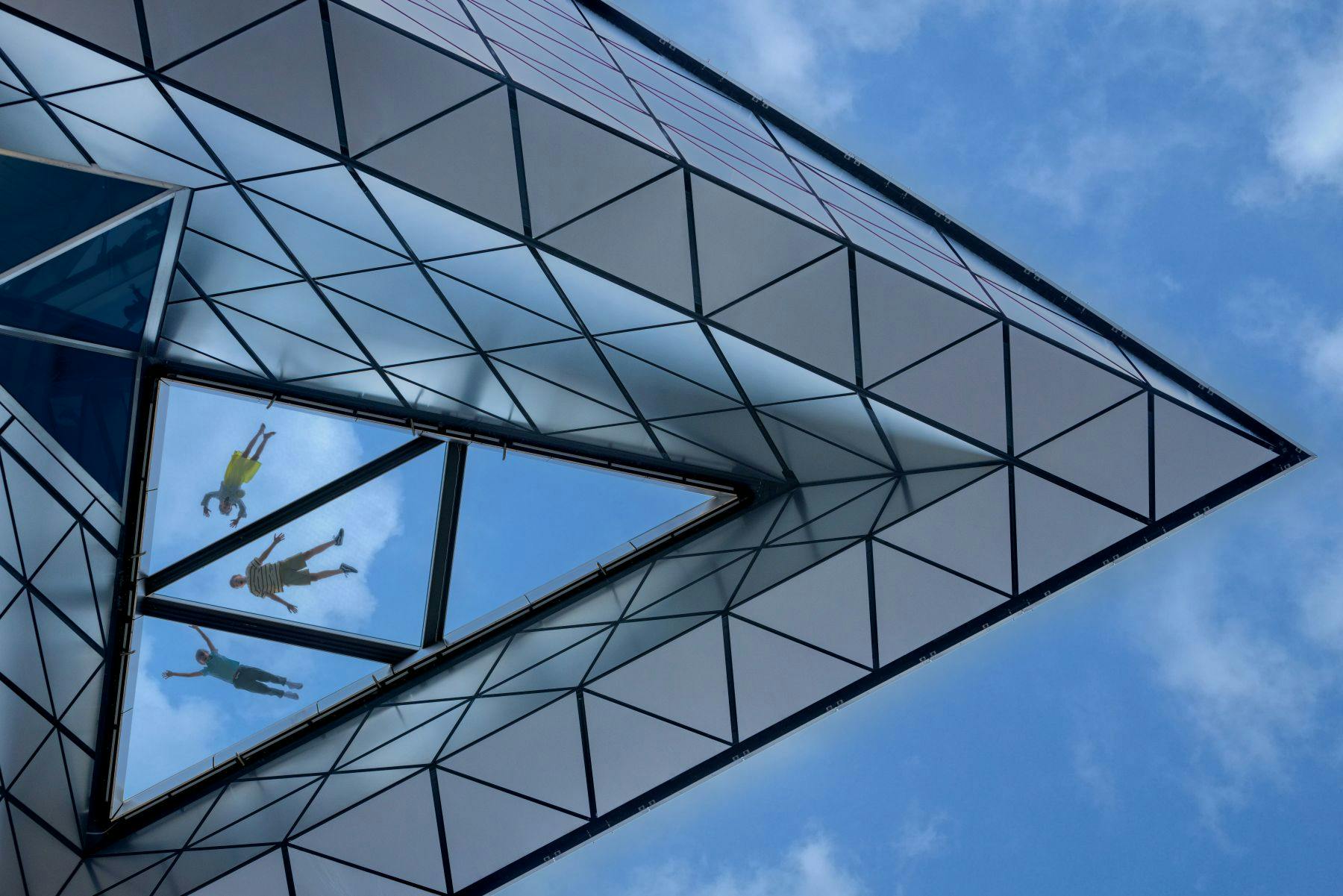 Un moderno techo de cristal con motivos triangulares, que refleja el cielo y muestra una figura vista a través de una sección transparente.