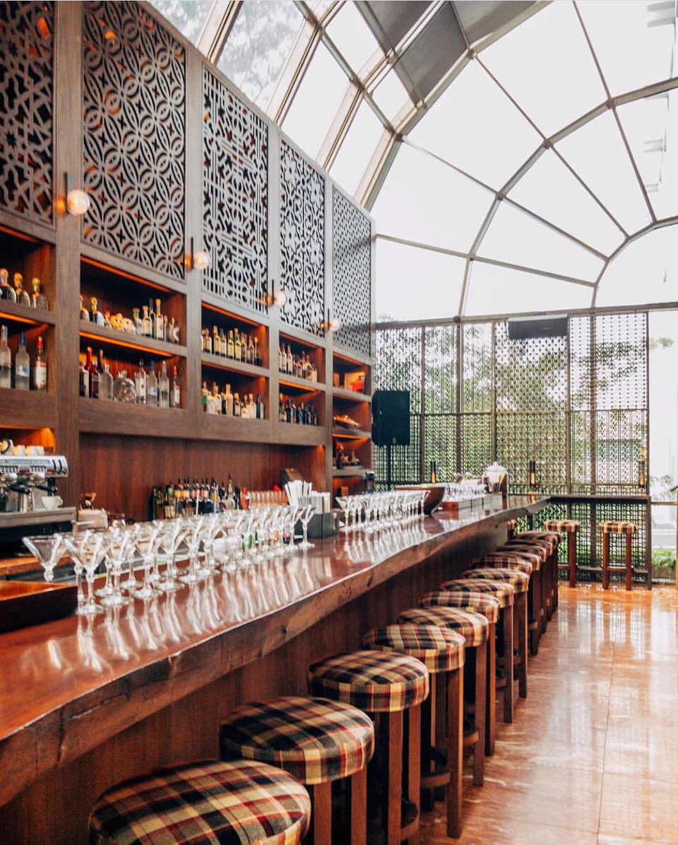 Spacious bar with a long wooden counter, rows of glassware, plaid upholstered stools, and liquor shelves under an arched glass ceiling.