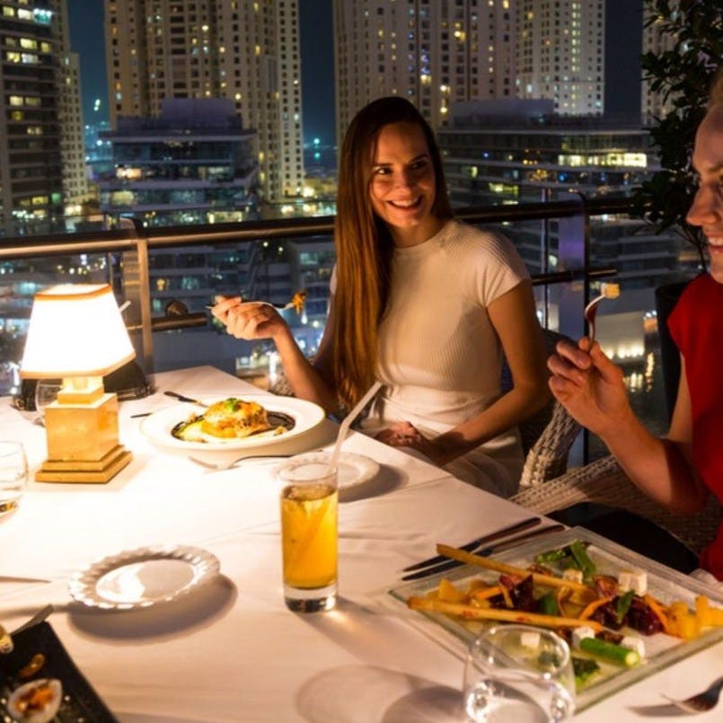 Dvě ženy večeří v dobře osvětlené venkovní restauraci s panoramatickým výhledem na město a výškovými budovami, které jsou v noci vidět v pozadí.