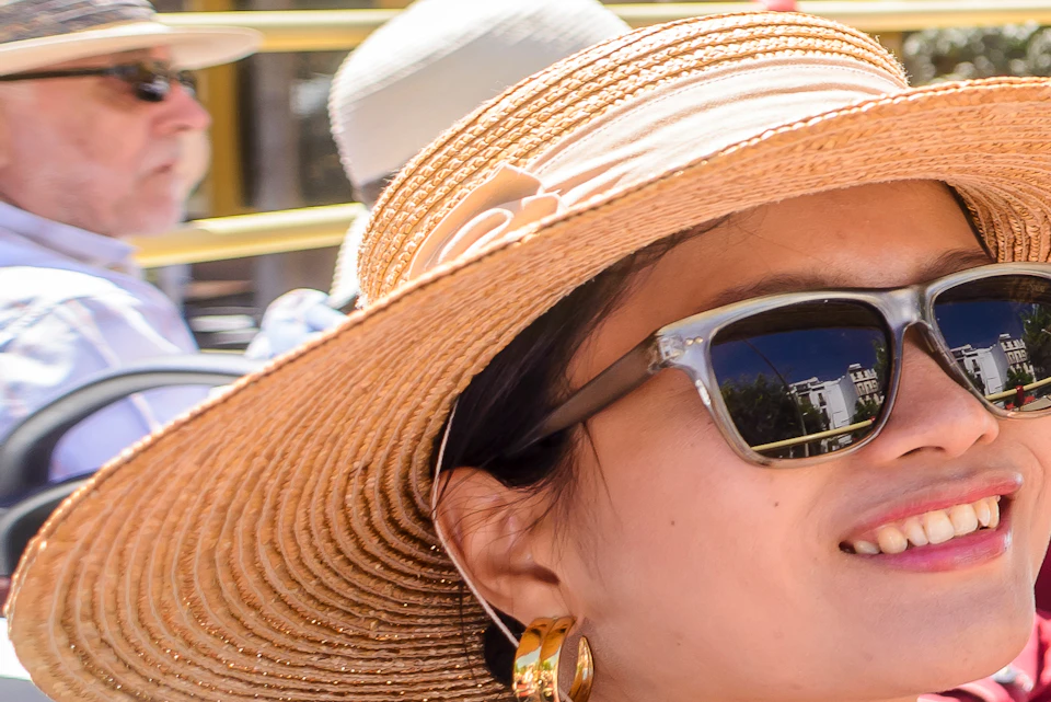 A smiling woman in a summer hat and sunglasses holds a brochure on an open-top bus tour in a city with historic buildings.