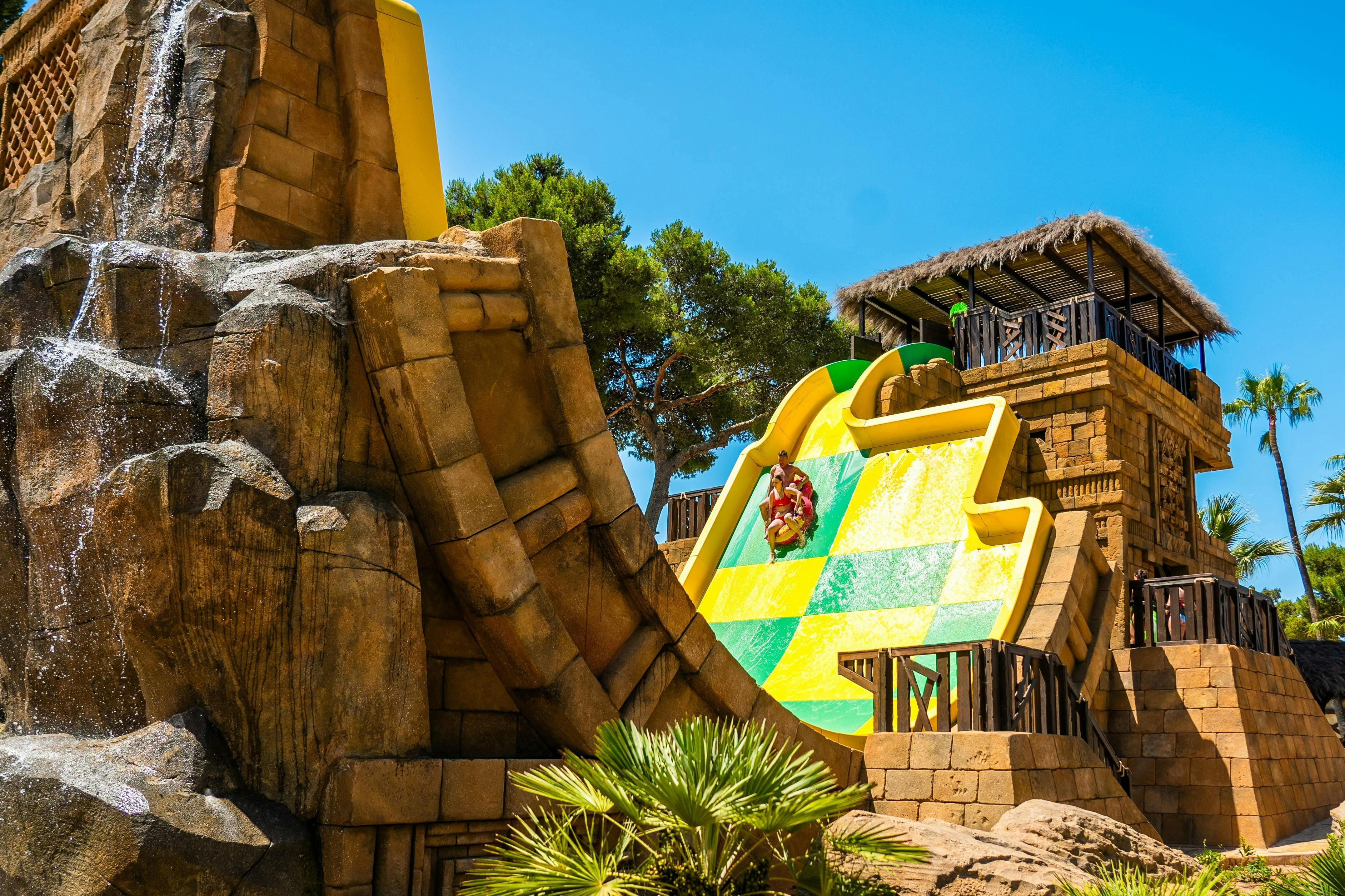 A large water slide with a green and yellow checkered pattern. A person is sliding down it. Surrounded by rocks and trees.