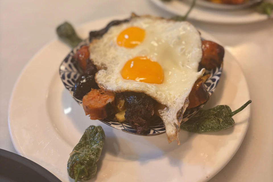 A plate with two sunny-side-up eggs atop mixed sausages and roasted vegetables, including green peppers.
