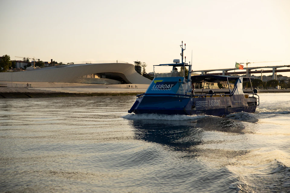 A blue boat labelled "LISBOAT" moves across calm water with a modern white building and a bridge in the background.