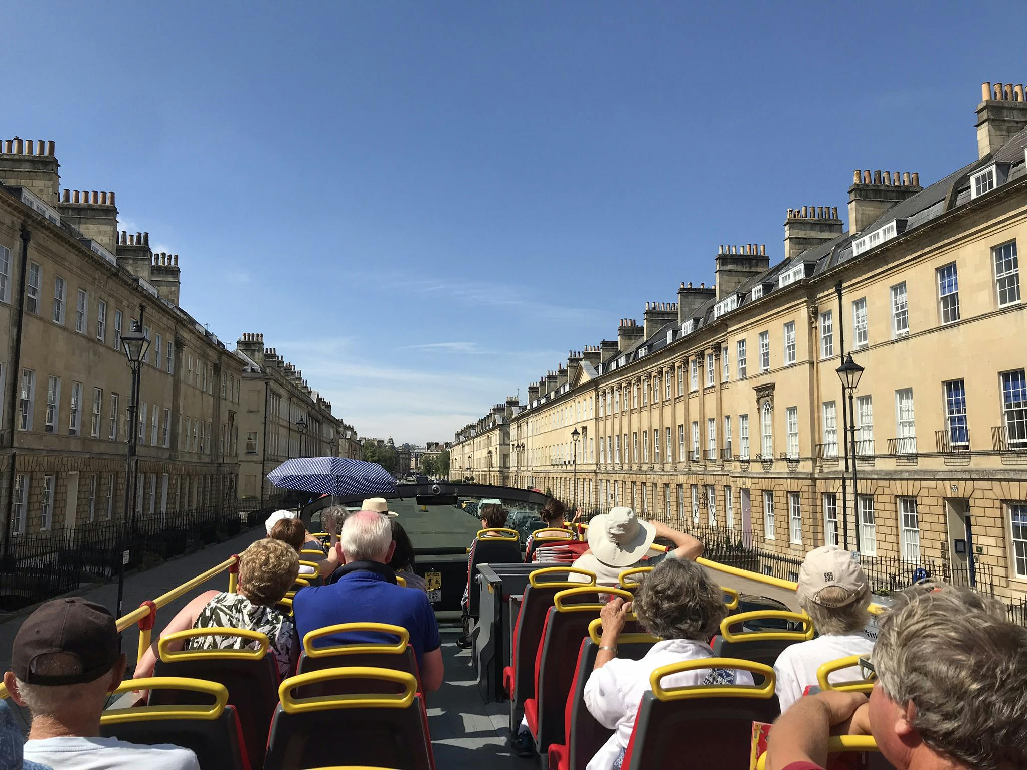 Οι τουρίστες κάθονται στο πάνω κατάστρωμα ενός διώροφου λεωφορείου μια ηλιόλουστη μέρα, βλέποντας σειρές ιστορικών κτιρίων κατά μήκος ενός δρόμου.