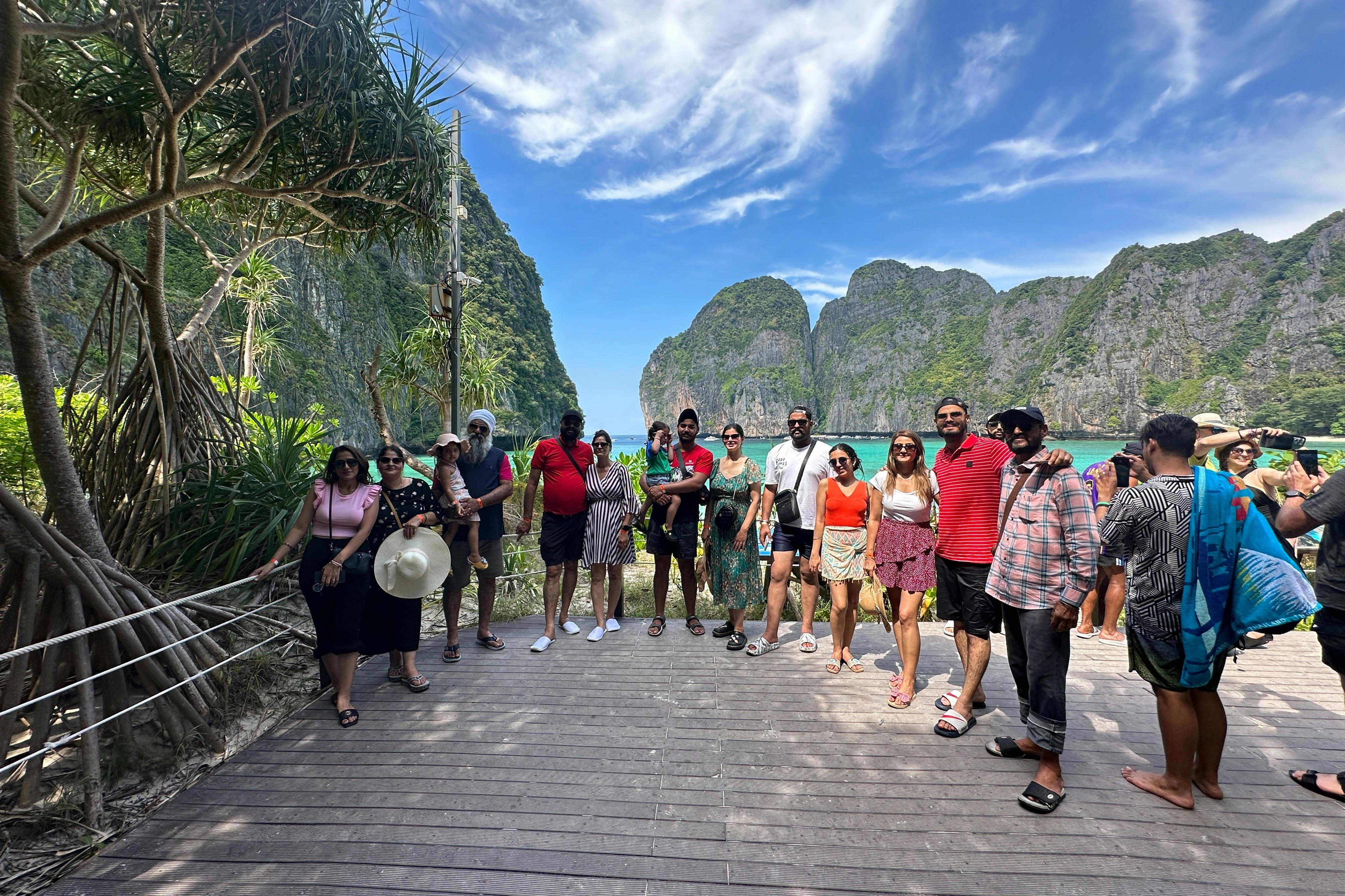 Μια ομάδα ανθρώπων στέκεται σε ένα ξύλινο κατάστρωμα με ένα γραφικό σκηνικό από γκρεμούς και γαλάζιο ουρανό, χαμογελώντας και ποζάροντας για μια φωτογραφία.