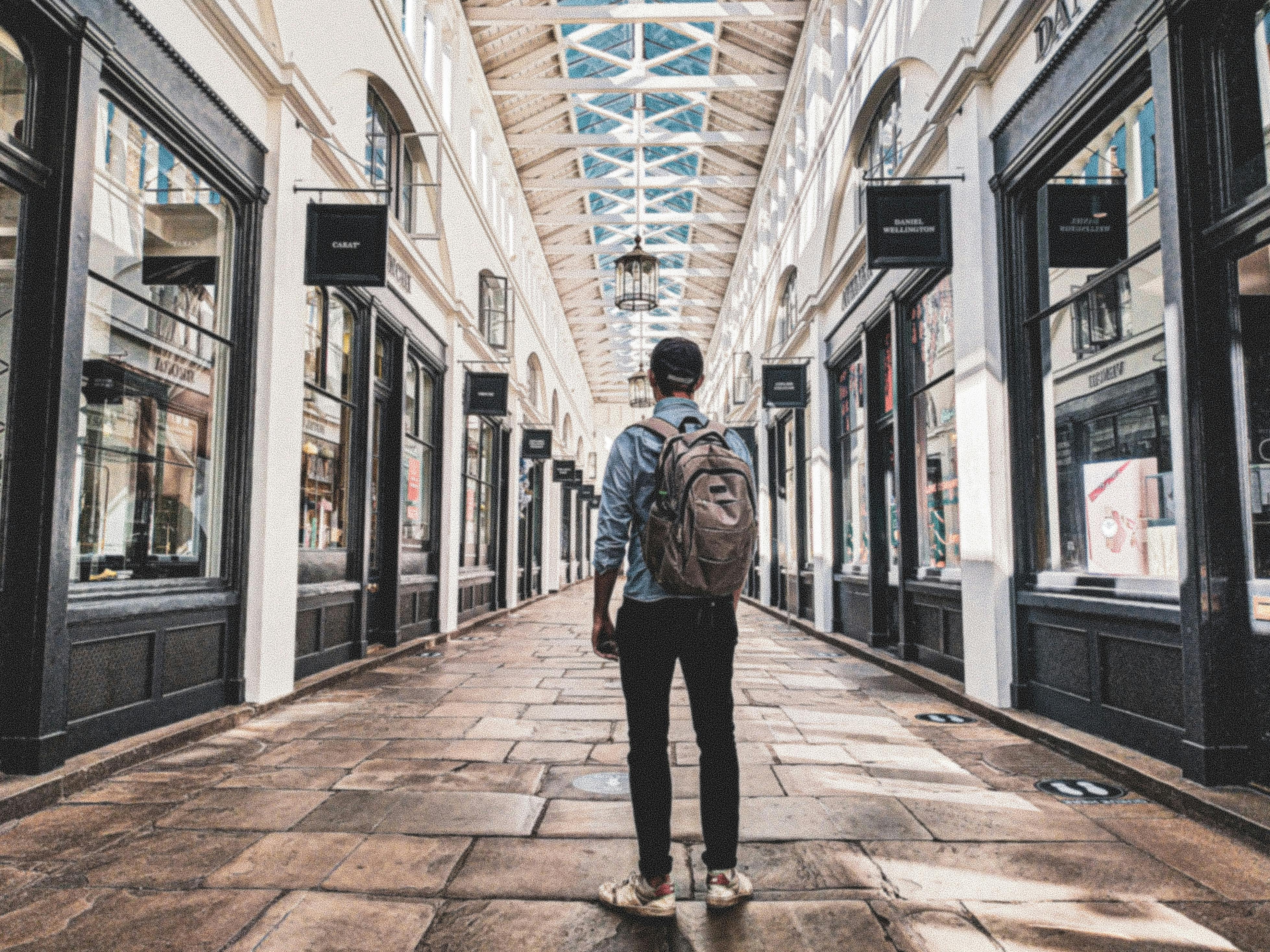 Person with a backpack walking through an indoor shopping arcade with glass ceiling and decorative lanterns.