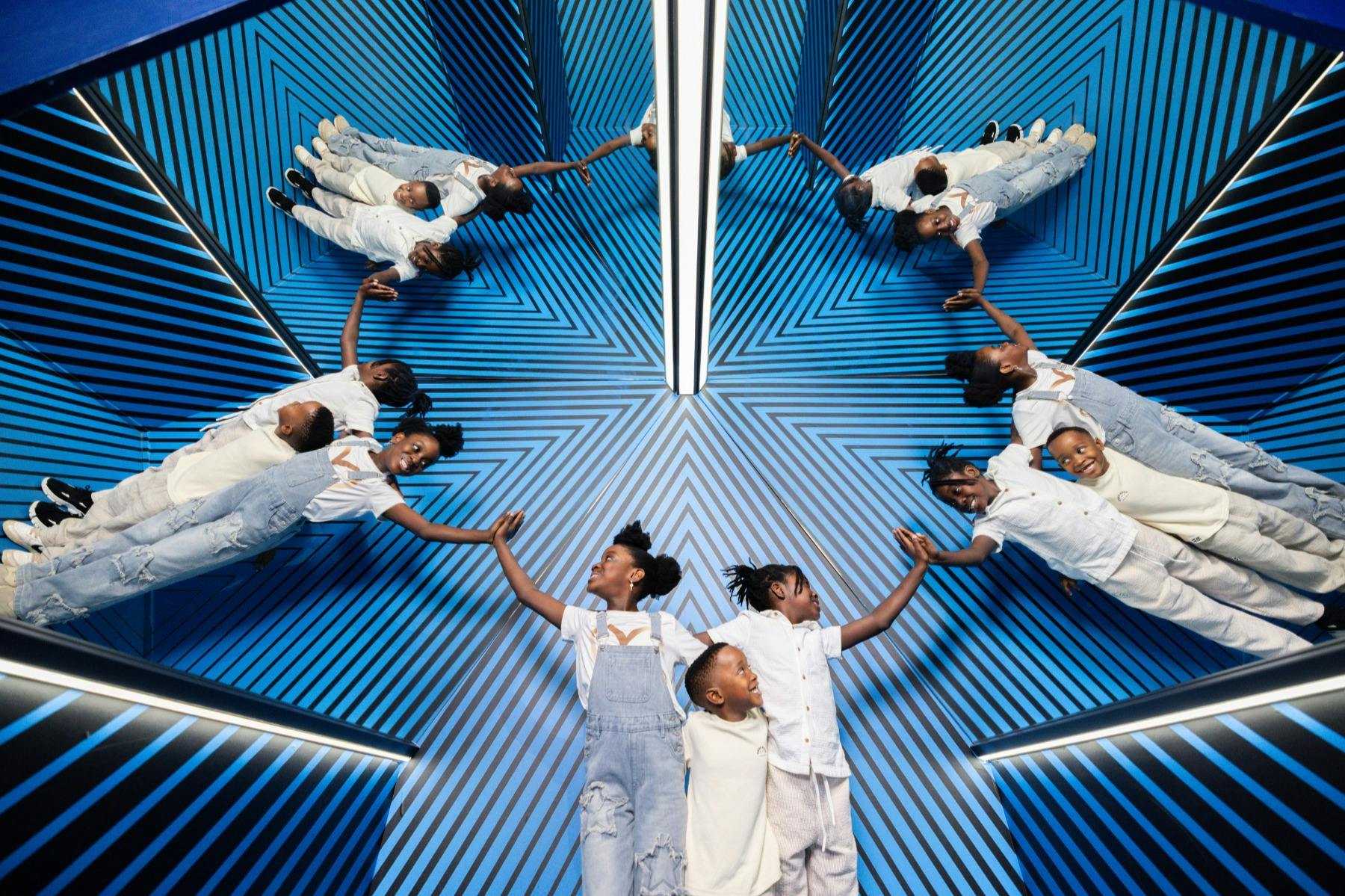 En grupp barn i lätta kläder poserar i ett rum med blårandiga väggar och speglar som skapar kalejdoskopiska reflektioner.