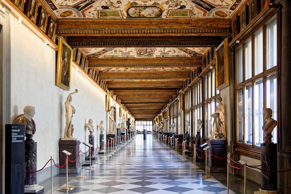 An ornate museum hallway with classical sculptures, framed paintings, and a painted ceiling, lined with checkered floor tiles.
