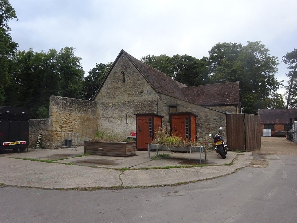 石头建筑，有两扇木门，附近停着一辆摩托车，前面有花盆。 后面有树木和另一栋建筑。