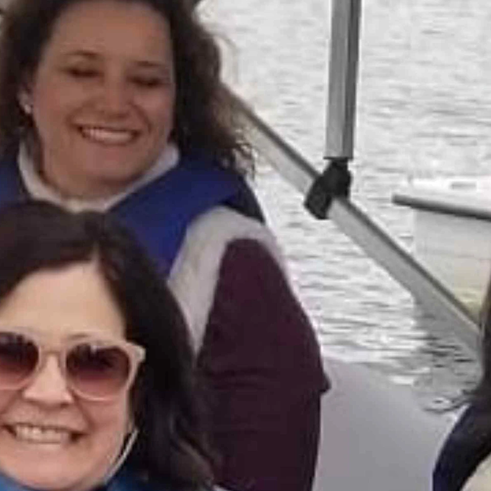 Four women, wearing life jackets, smile while sitting in a swan-shaped paddle boat at a dock with other boats in the background.
