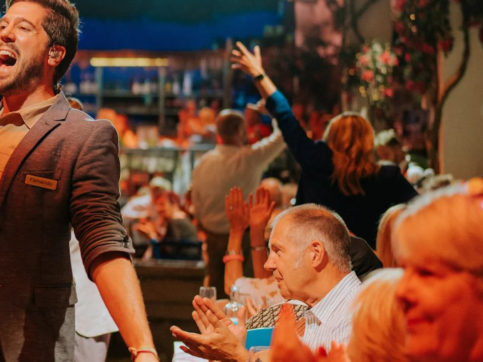 A lively scene of people dining and celebrating in a festive, well-lit venue with a performer engaging the crowd.