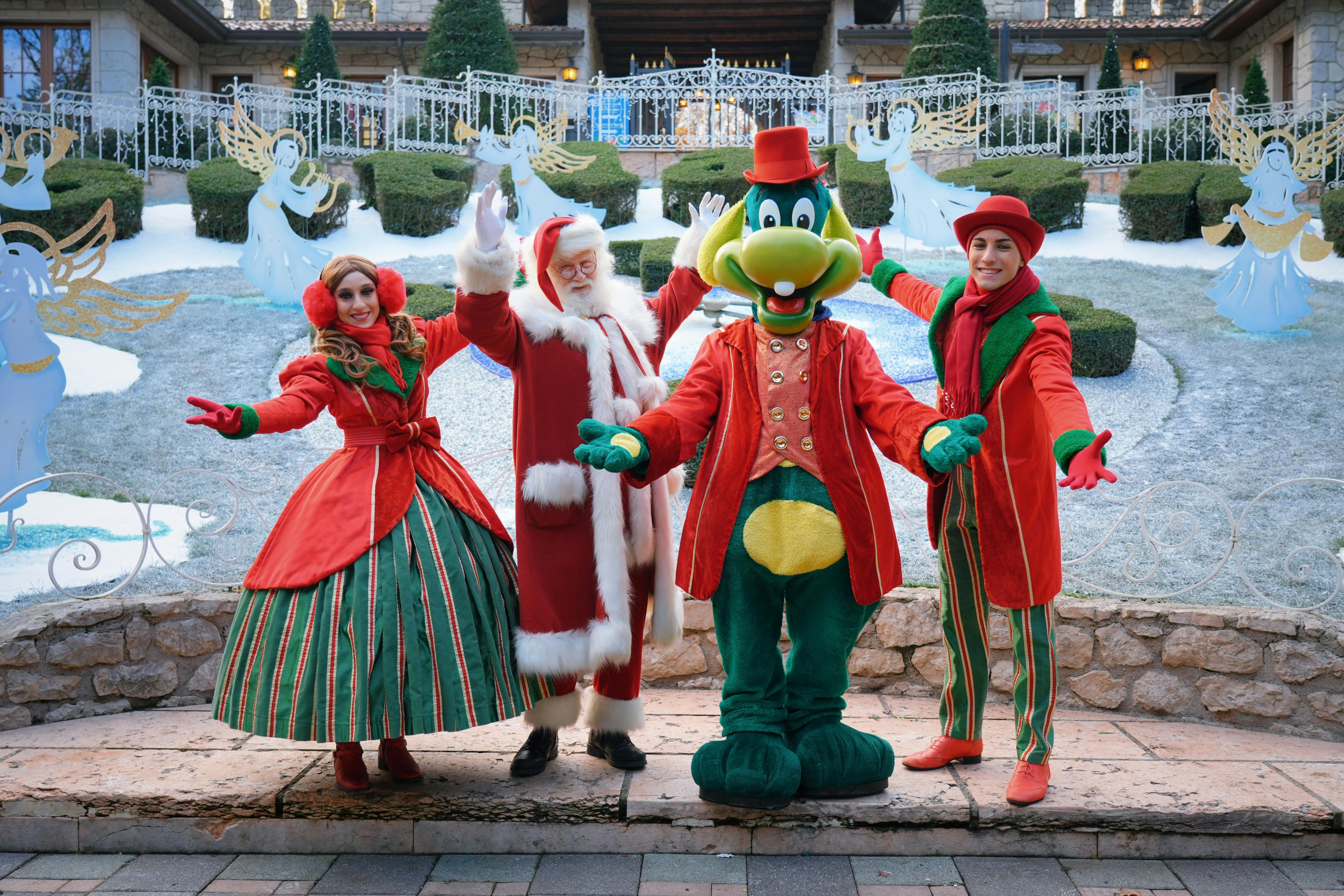 Člověk oblečený jako Santa, usměvavý kreslený maskot a elf stojí s otevřenou náručí před slavnostními zimními dekoracemi.