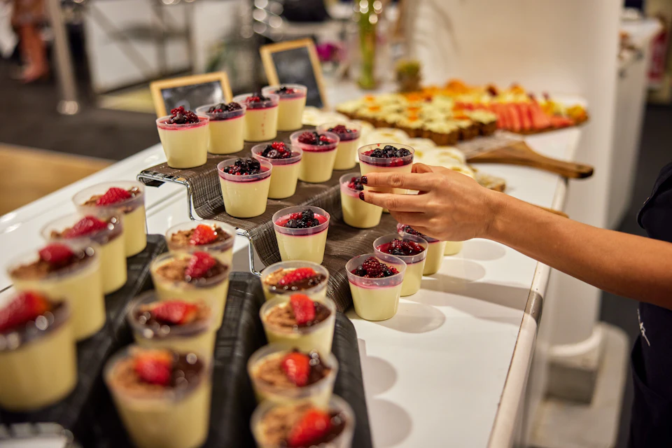 A hand reaching for a dessert cup topped with berries, among multiple identical cups on a serving counter.