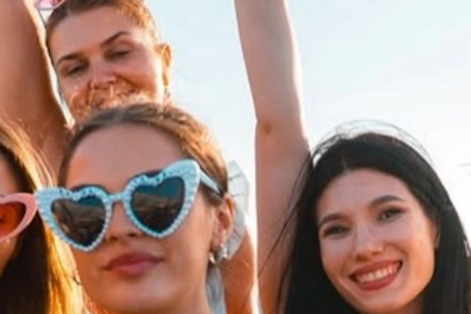 A group of women outdoors wearing colorful outfits and heart-shaped sunglasses, smiling and enjoying sunny weather.