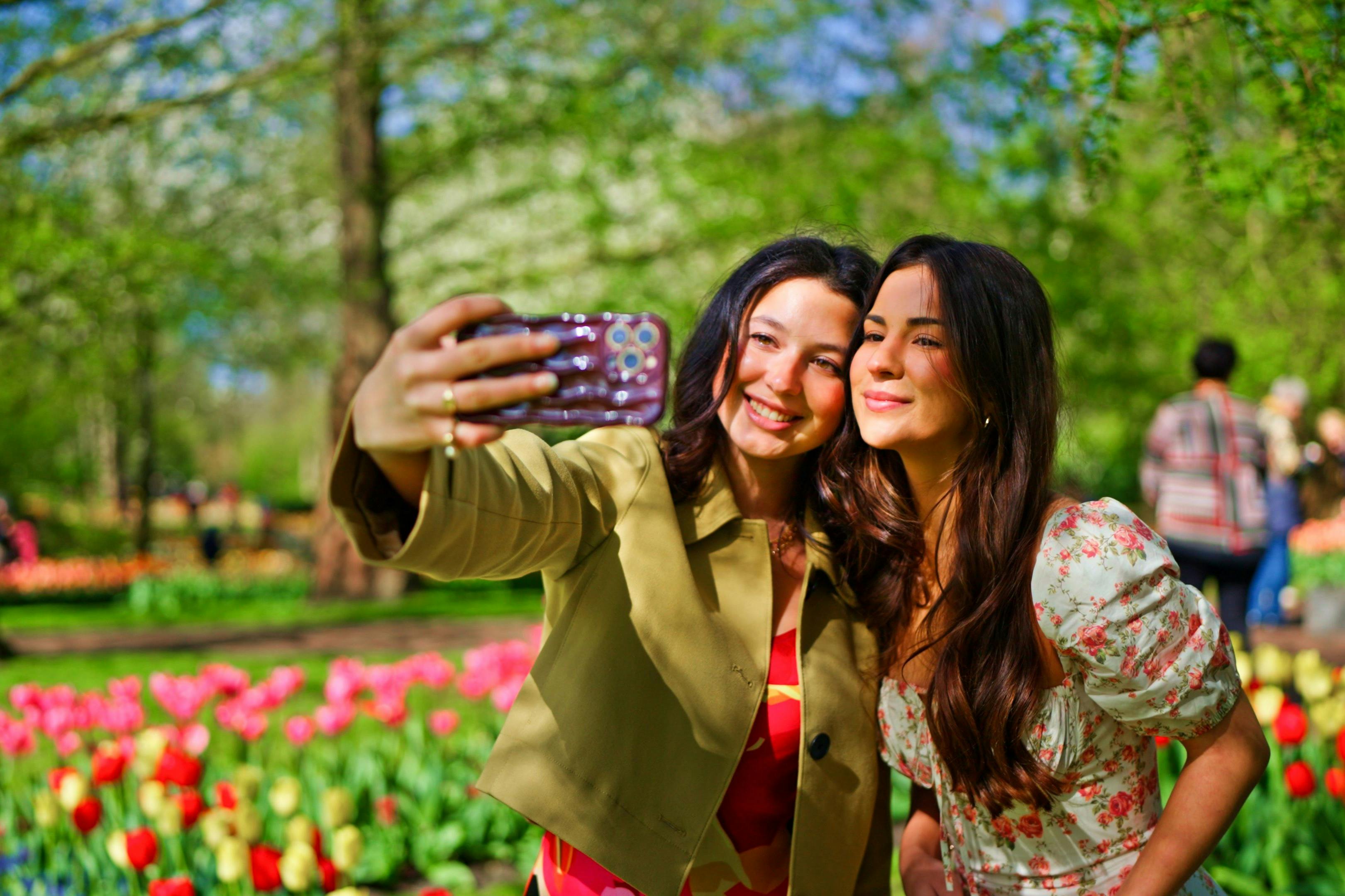 Two women posing for a selfie in a garden with blooming flowers and greenery in the background on a sunny day.