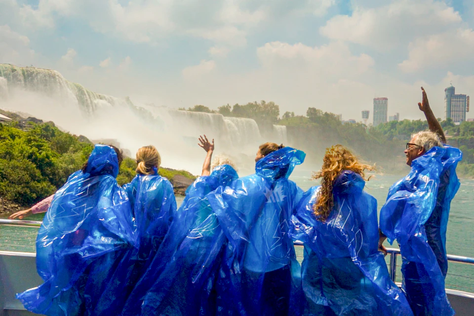 Group of people wearing blue ponchos, viewed from behind, facing a large waterfall under a cloudy sky.