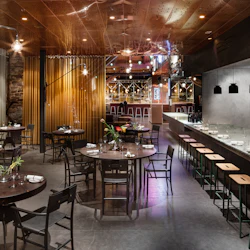 Modern restaurang med runda bord, uppsättning med servetter och glas, en lång bardisk med pallar, taklampor och blomsterarrangemang.