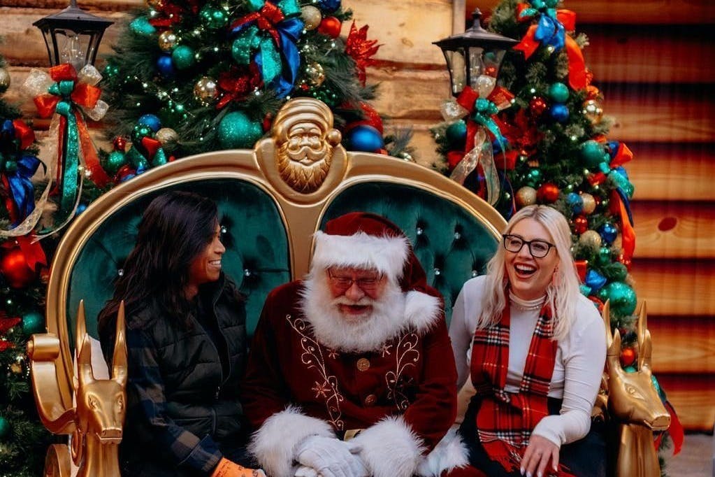 두 명의 여성이 산타클로스 양쪽에 기쁘게 앉아 있습니다. 산타클로스는 장식과 활로 장식된 크리스마스 트리 앞에 앉아 있습니다.