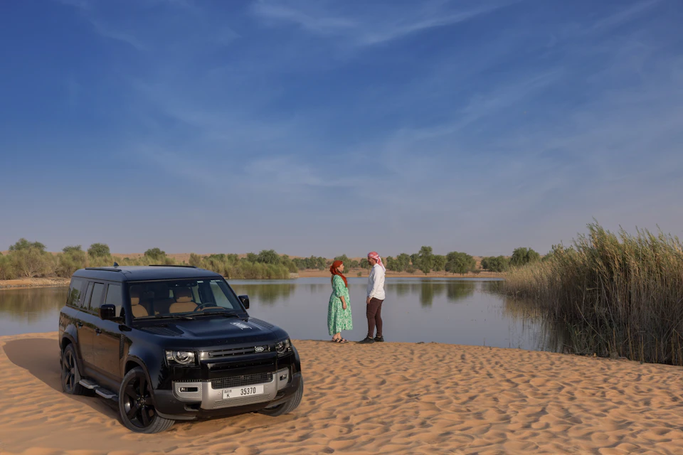 A black SUV parked on sand beside two people standing near a calm waterbody under a clear blue sky.