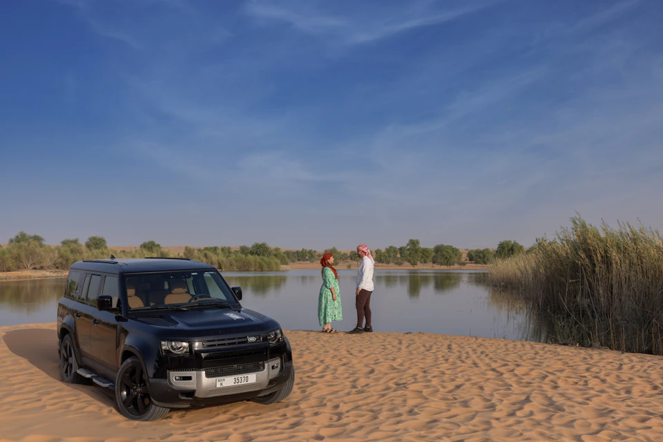 A black SUV parked on sand beside two people standing near a calm waterbody under a clear blue sky.