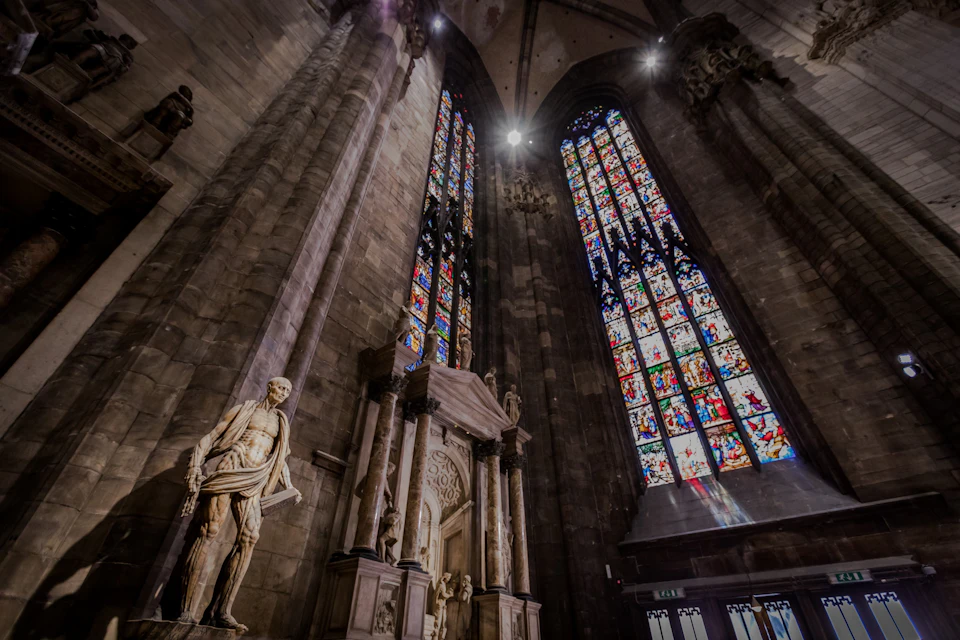 Interior of a cathedral with large stained glass windows and statues, including a prominent marble figure on the left.